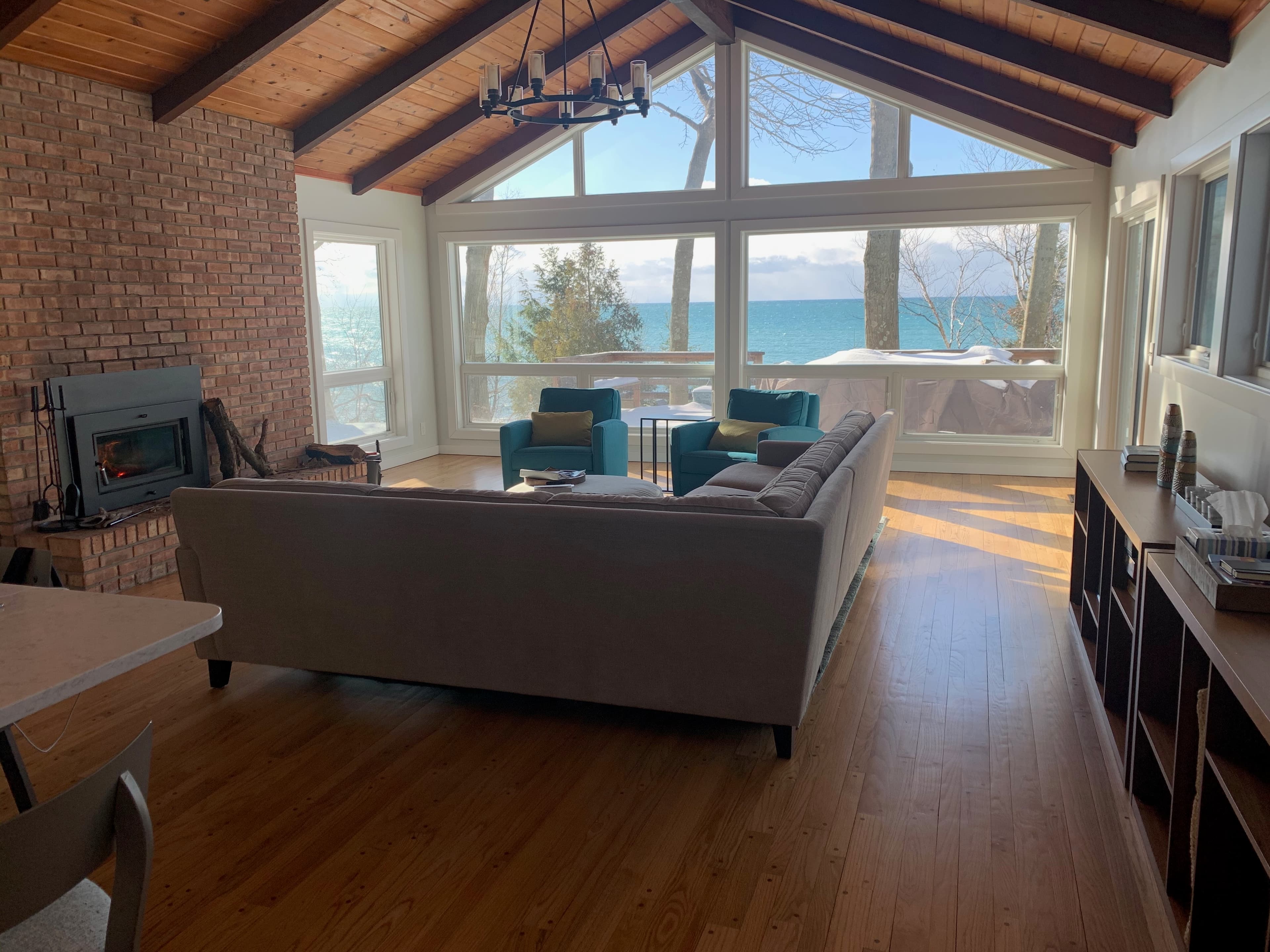 Open-concept living room with vaulted wood ceiling, brick fireplace, and floor-to-ceiling windows overlooking Lake Michigan